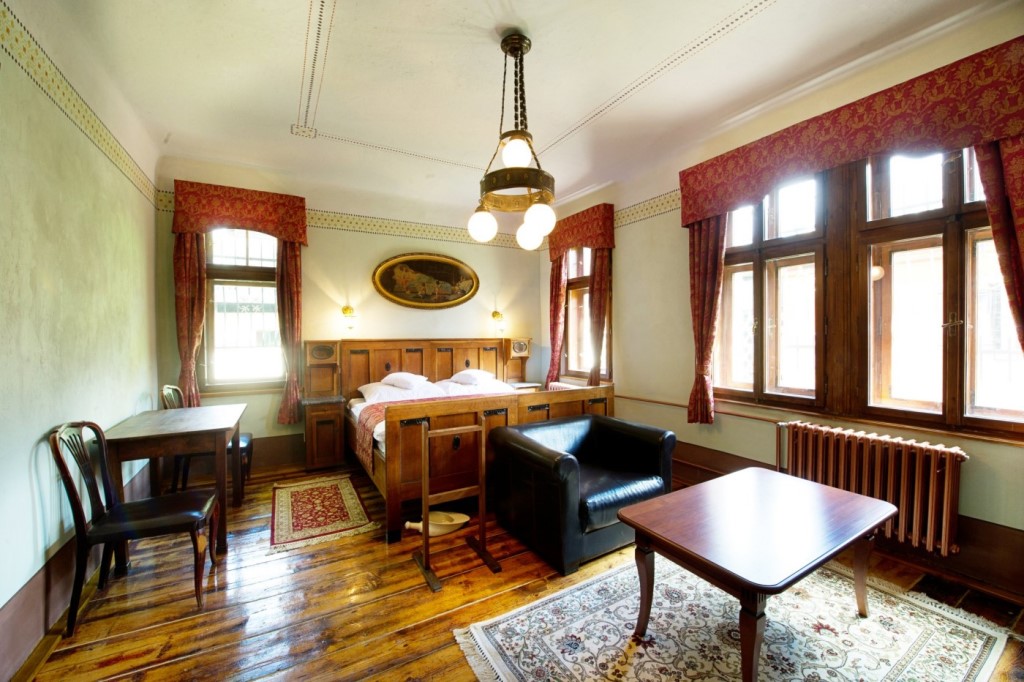 Pokoj Vlasta Burian - prostorný pokoj s dřevěným nábytkem zdobeným kovovými prvky stojícím na původní dřevěné podlaze.